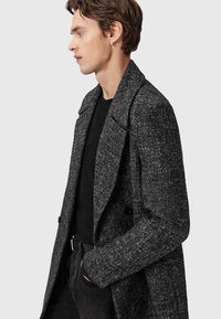 Grå tekstureret frakkemed krave og dobbeltradet design. Har et subtilt mønster og bæres over en sort sweater.