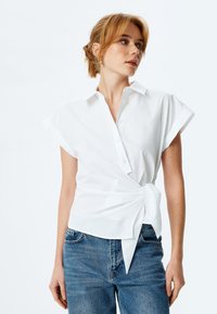 Camicia bianca a maniche corte con bottoni, dotata di design avvolgente e nastro legato in vita, realizzata in morbido tessuto di cotone. Abbinata a jeans di denim.
