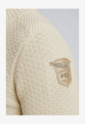Beiger Strickpullover mit Rippkragen, der ein strukturiertes Muster und einen Aufnäher mit detaillierter Bestickung am Ärmel aufweist.