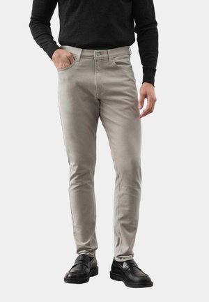 Mand iført beige slim-fit bukser, sort langærmet top og sorte lædersko, med den ene hånd i lommen og den anden ved siden.