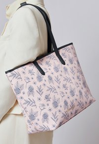 Person in cremefarbener Kleidung trägt eine rosa Tragetasche mit schwarzem Floral-Muster und schwarzen Griffen über der Schulter.