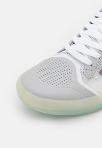 Šedá atletická obuv se strukturovaným koženým svrškem, síťovinovými detaily, perforovanou špičkou, bílými tkaničkami a průhlednou podrážkou.