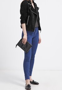 Chaqueta de motorista de ante negro, blusa de lunares, jeans ajustados azules, sosteniendo un clutch negro. Mocasines oscuros con un acabado brillante. Diseño minimalista.