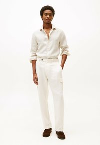 Camisa de lino beige claro con mangas largas, con un sutil patrón de rayas, combinada con pantalones de lino blanco roto y zapatos marrones.