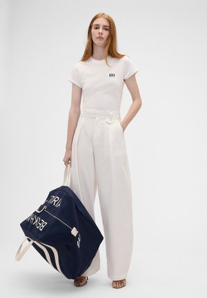 Junge Frau in weißem GAP-T-Shirt und weit geschnittenen Hosen hält eine große dunkelblaue Duffel-Tasche mit weißen Trägern und Text.
