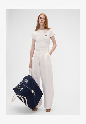Jeune femme en t-shirt blanc GAP et pantalon à jambes larges tenant un grand sac de sport bleu marine avec des anses blanches et un texte.