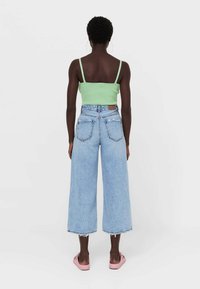 Top cropped verde aderente, jeans a vita alta azzurri chiari a gamba larga con orlo sfilacciato e scarpe rosa slip-on. Modello visto da dietro.