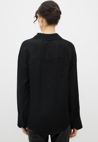 Maglietta nera a maniche lunghe con un motivo sottile, dotata di colletto e polsini con bottoni. Tessuto liscio, vestibilità rilassata che si estende fino ai fianchi.