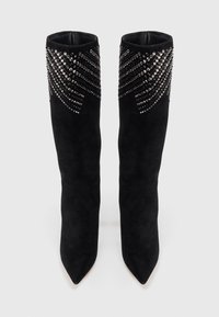 Botas altas de ante negro hasta la rodilla con punteras afiladas, decoradas con tachuelas metálicas plateadas en un patrón radiante cerca del borde superior.