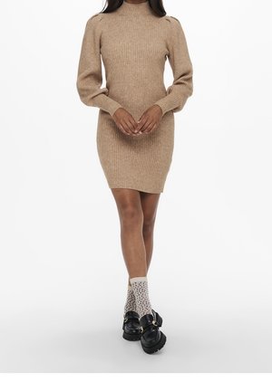 Vrouw draagt een beige geribbelde gebreide jurk met lange ballonmouwen, patroon enkelsokken en chunky zwarte loafers, staand tegen een witte achtergrond.