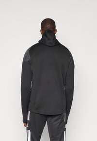 Schwarzer Pullover-Hoodie mit hohem Kragen und tailliertem Design. Verfügt über glattes Material mit glänzenden schwarzen Schulterakzenten und Daumenlöchern in den Ärmeln.