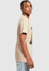 Beige bomulds t-shirt med korte �ærmer og rund halsudskæring, med et grafisk tryk i mørke farver på forsiden.