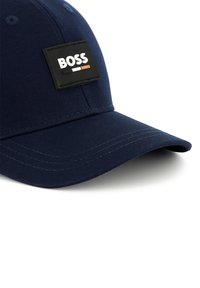 Marineblauwe pet met een platte rand, voorzien van een zwart rechthoekig label met het woord "BOSS" in witte, oranje en zwarte letters.