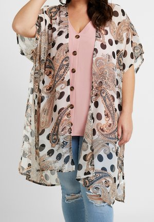 Frau trägt ein durchsichtiges Kimono mit Paisley- und Polka-Dot-Muster über einem rosa geknöpften Oberteil und zerrissenen hellblauen Jeans, Hand in Kopfnähe erhoben.