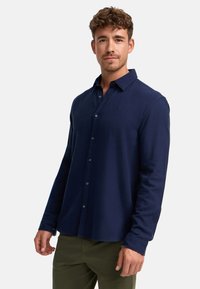 Camisa de botones azul marino, de manga larga, hecha de una tela texturizada. Presenta un cuello y un logo sutil en el pecho. Incluye puños con botones.