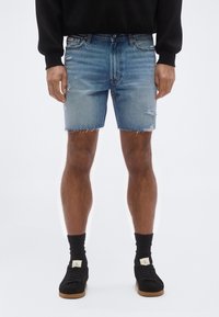 Άνδρας που φοράει ανοιχτό μπλε ξεβαμμένα τζιν σορτς, μαύρες κάλτσες και μαύρα αθλητικά παπούτσια με καφέ σόλες, στέκεται μπροστά σε απλό λευκό φόντο.