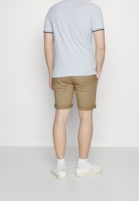 Světle modré tričko s tmavým lemem, khaki šortky s ohrnutými nohavicemi a bílé tenisky na osobě pohledem zepředu proti jednobarevnému pozadí.