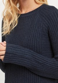 Pull en maille bleu marine présentant une texture côtelée, un col rond et des poignets allongés pour plus de chaleur. Tissu doux et confortable avec une coupe ajustée.