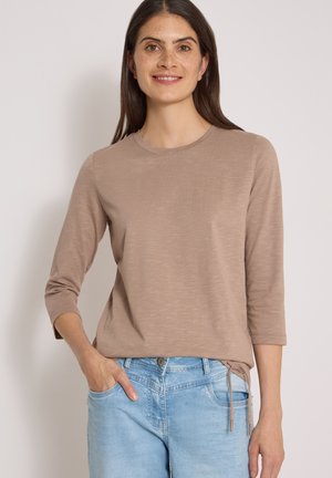 Femme aux longs cheveux foncés souriante, portant un haut beige à manches trois-quarts et un jean bleu clair, main dans la poche, debout devant un fond uni.