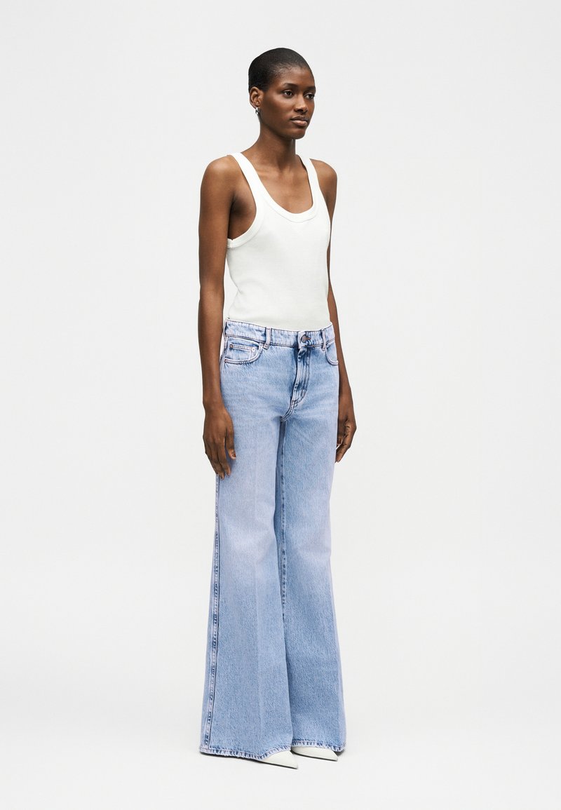 Vrouw staand, gekleed in een witte mouwloze tanktop, lichtblauwe high-waisted wijde jeans en witte puntige schoenen, tegen een effen achtergrond.