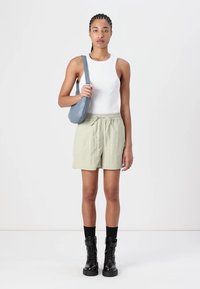 Bijela majica bez rukava, svijetlozelene kratke hlače, siva crossbody torba, crne čizme do koljena. Model stoji pred jednostavnom pozadinom.