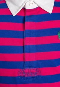 Bomullsblandad pikétröja med horisontella rosa och blå ränder, vit krage, fyraknappsknut och grön broderad logotyp på vänster sida.