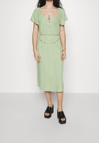 Vaaleanvihreä midi-mekko, jossa on lyhyet puffihihat, etuvetoketju ja kultaketjuvyö. Yhdistetty mustien paksupohjaisten korkokenkien kanssa.