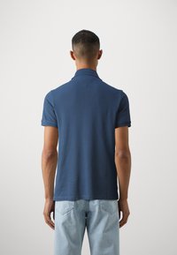 Marineblauwe polo met korte mouwen, geribbelde kraag en een gestructureerde afwerking. Gekoombineerd met lichtblauwe denim jeans, gezien vanaf de achterkant.