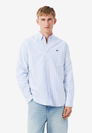 Joven con camisa abotonada de rayas verticales blancas y azul claro con un pequeño logo de cocodrilo verde y jeans azul claro.