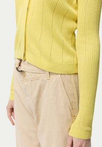Gul ribbad stickad tröja med långa ärmar och en kortad fåll, ihopparad med beiga byxor som har knapp- och bältesslingsdetaljer.