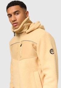 Chaqueta de forro polar beige con capucha, tejido texturizado, bolsillo con cremallera en la parte delantera, panel gris en el pecho y parche de la marca en la manga.