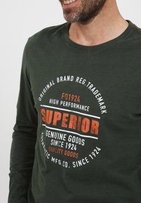 Langærmet, mørkegrøn bomulds skjorte med rund halsudskæring og trykt tekst i hvid og orange, herunder "SUPERIOR" og "GENUINE GOODS."