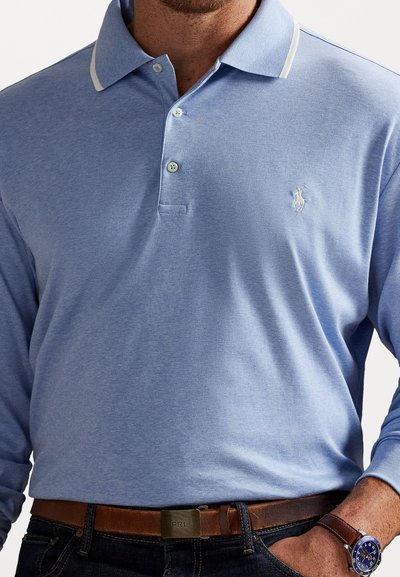 Cămașă polo albastră deschis cu mânecă lungă, cu guler cu dungi albe, trei butoane și un mic logo brodat pe piept. Purtați cu o curea maro.