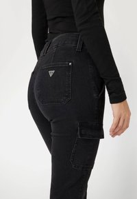 Pantalons en denim noir avec poches de style cargo, présentant un logo en métal en forme de triangle sur la poche arrière et une coupe ajustée. Texture lisse.