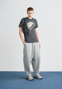 Siva majica z zlatnim logotipom, sive trenirke in sivi športni copati. Majhna črna torbica, nošena čez telo. Preprost dizajn, sproščen kroj. Modro ozadje preproge.