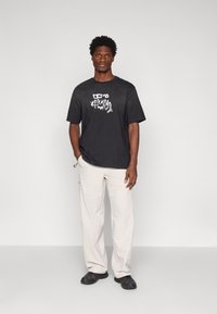 Czarny t-shirt graficzny z zniekształconym białym nadrukiem, w połączeniu z jasnobeżowymi spodniami cargo i czarnymi butami sportowymi. Prosty, luźny krój.