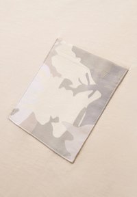 Tejido beige con un parche de camuflaje gris claro texturizado cosido en el lado izquierdo, con formas curvas y bordes cosidos.