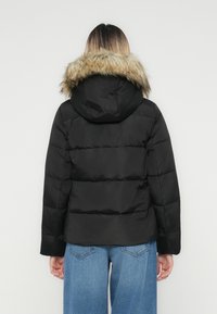 Personen vender væk iført en sort pufferjakke med en hætte trimmet med pels og blå jeans mod en ensfarvet hvid baggrund.