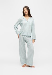 Femme debout pieds nus portant un haut de pyjama ample à rayures bleu clair et blanches, boutonné, à manches longues, et un pantalon large assorti sur fond blanc.