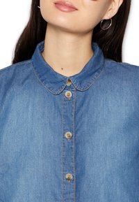 Camicia in denim con colletto a punta, chiusura a bottoni e cuciture a contrasto. Colore azzurro chiaro con una texture liscia e quattro bottoni.