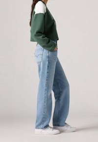 Persona vestida con un suéter corto verde y blanco, jeans de tiro alto azul claro, zapatillas blancas, de pie con las manos en los bolsillos.
