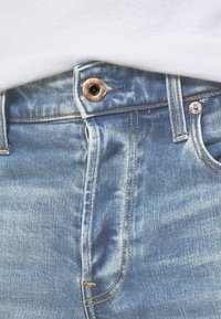 Close-up da cintura de um par de jeans de ganga azul, com um botão de cobre visível e costura laranja, sobre um fundo de tecido branco.