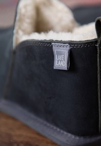 Black suede slipper with a fuzzy cream lining, featuring a gray stitching and a tag labeled "LAKELAND." Soft texture and rounded shape.