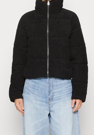 Personne portant une doudoune matelassée noire avec un col montant et une fermeture éclair argentée, associée à un jean bleu clair taille haute.