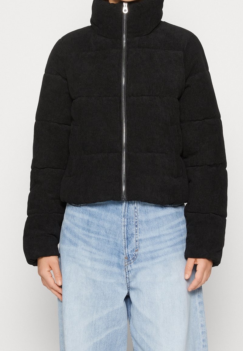 Personne portant une doudoune matelassée noire avec un col montant et une fermeture éclair argentée, associée à un jean bleu clair taille haute.