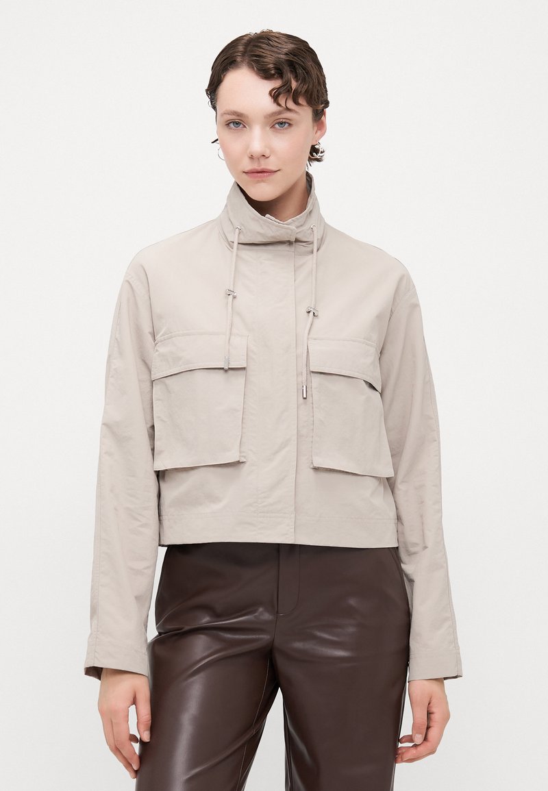 Beige cropped jacket with high collar, two large front pockets, drawstring closure, and long sleeves. Smooth, lightweight fabric texture.