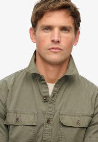 Camisa de algodón verde oliva con cuello alto, dos bolsillos en el pecho y botones marrones. Tejido texturizado con un corte relajado y un diseño casual.