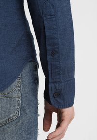 Tmavě modrá košile s dlouhým rukávem vyrobená z texturované tkaniny, se zapínacími manžetami a zaobleným lemem, nošena s vybledlými modrými džíny.