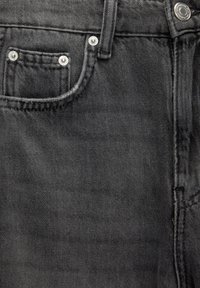 Gros plan sur un jean denim noir montrant la poche avant, la passants de ceinture, les coutures et les rivets métalliques aux coins de la poche ainsi que le bouton de la ceinture.