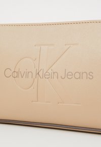 Beige lompakko, jossa on kohokuvioitu "CK" ja "Calvin Klein Jeans" -teksti, neulotut reunat ja sileä pinta.